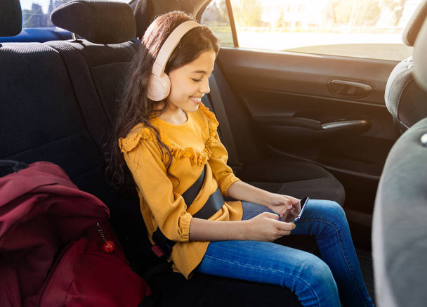 Joyeux jeune fille avec casque bénéficie de son smartphone tout en étant assis dans le siège arrière des voitures, la lumière du soleil en streaming dans, sac à dos de l'école rouge à côté d'elle - Photo, image