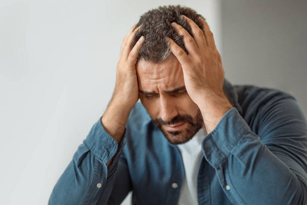 Nieszczęśliwy facet w średnim wieku wykazujący dyskomfort i ból głowy, dotykając głowy doświadcza depresji i stresu siedząc w domu. Problemy zdrowotne i cierpienie emocjonalne - Zdjęcie, obraz