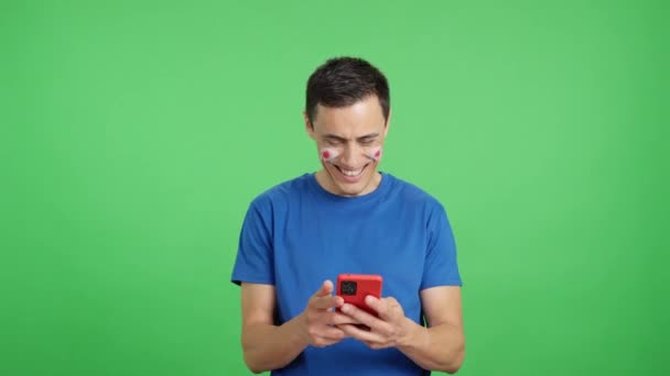 Vídeo de estudio con fondo de croma de un partidario de Japón con la bandera de Japón pintada en su cara, mirando su teléfono móvil sonriendo y mostrándolo a la cámara con la pantalla en croma key - Imágenes, Vídeo