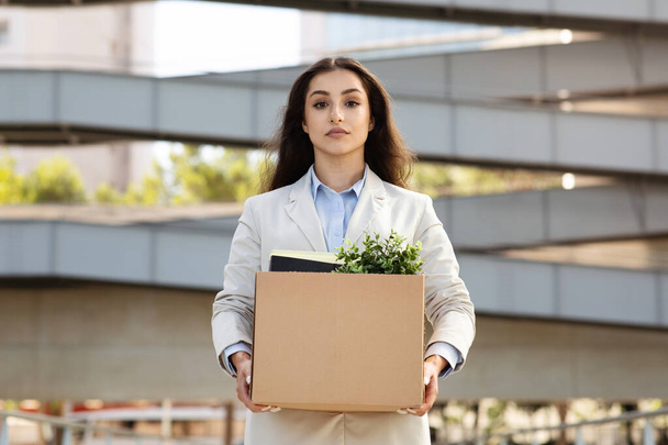 Sad serious young caucasian woman manager hold cardboard box filled with office essentials, green potted plant, in city office, outdoors. Problems with work, dismissal, quit - Photo, Image