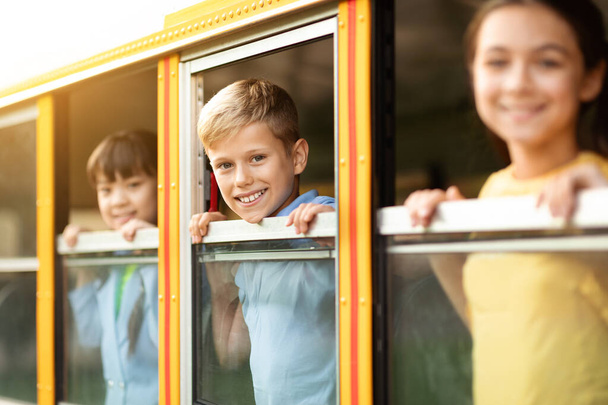 Enfants heureux regardant par la fenêtre du bus scolaire jaune, leurs visages joyeux tournés vers la caméra, groupe d'élèves appréciant le voyage, prêt pour le voyage ensemble, accent sélectif sur le garçon - Photo, image