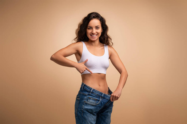 Happy attractive brunette slender millennial woman wearing huge jeans, showing results of diet, demonstrating loose in waist, beige studio background, slimming concept, copy space - Photo, Image