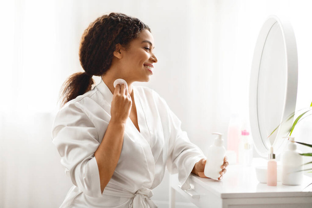 Belle femme noire avec des tampons de coton nettoyant la peau du cou à la maison, attrayante jeune femme afro-américaine assise à la coiffeuse et regardant dans le miroir, faisant routine quotidienne de beauté - Photo, image