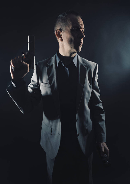 Spion in elegant pak met pistool pistool in donkere studio shot foto met effen achtergrond en kopieer ruimte. - Foto, afbeelding