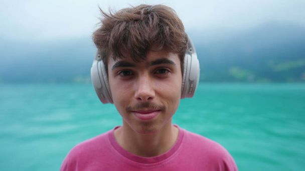 Portrait of young boy putting headphones listening to audio overlooking lake shire landscape. Handsome teenager engaged with music, podcast, or audiobook looking at camera - Photo, Image