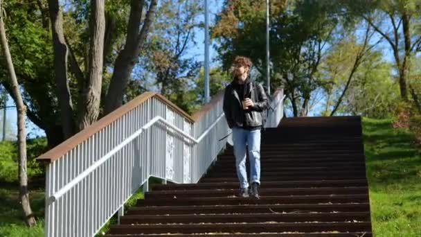 Uśmiechnięty stylowy facet z plecakiem cieszący się spacerkiem po parku podczas picia kawy. Styl życia. Koncepcja jesiennych spacerów. Koncepcja czasu rzeczywistego. - Materiał filmowy, wideo