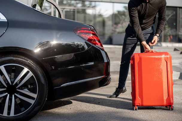 Ο Σοφέρ βάζει κόκκινη βαλίτσα σε ένα πορτ-μπαγκάζ αυτοκινήτου, από κοντά. Έννοια επαγγελματικών ταξιδιών, προσωπικού οδηγού ή ταξί πολυτελείας - Φωτογραφία, εικόνα