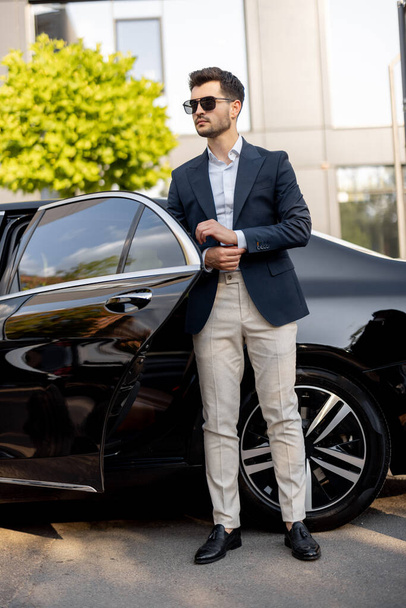 Πορτρέτο ενός όμορφου επιχειρηματία που στέκεται κοντά σε πολυτελές αυτοκίνητο σε εξωτερικούς χώρους. Ένας κομψός άνθρωπος βγαίνει από ένα premium αυτοκίνητο - Φωτογραφία, εικόνα