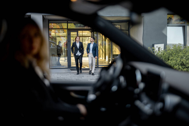 Deux hommes d'affaires marchent vers la voiture à partir d'un bâtiment, vue à travers une porte de véhicule. Le conducteur les attend dans la voiture. Concept de transport d'affaires - Photo, image