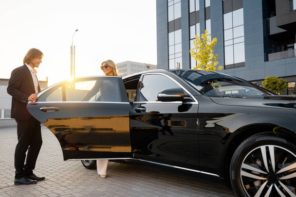 Деловой человек помогает деловой леди сесть в машину, открывая дверь машины возле офисного здания на закате. Концепция делового образа жизни и транспорта - Фото, изображение
