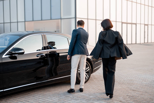 Businessman and businesswoman walk together to car on parking lot near modern building, going to drive by luxury car. Man opens vehicle door - Photo, Image