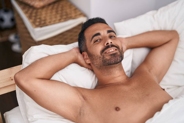 Junger hispanischer Mann lächelt selbstbewusst auf Bett im Schlafzimmer liegend - Foto, Bild