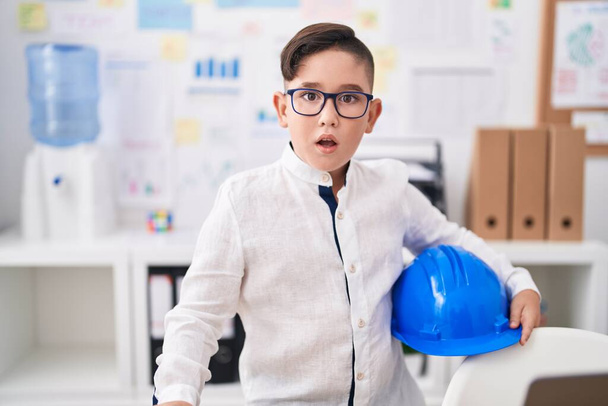 Genç İspanyol çocuk ofiste mimar şapkası tutuyor. Korkmuş ve ağzı açık kalmış, şaşırmış ve şaşırmış.  - Fotoğraf, Görsel