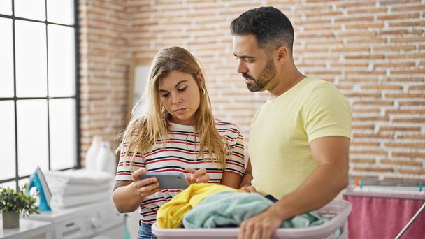 Man and woman couple holding basket with clothes using smartphone at laundry room - Photo, Image