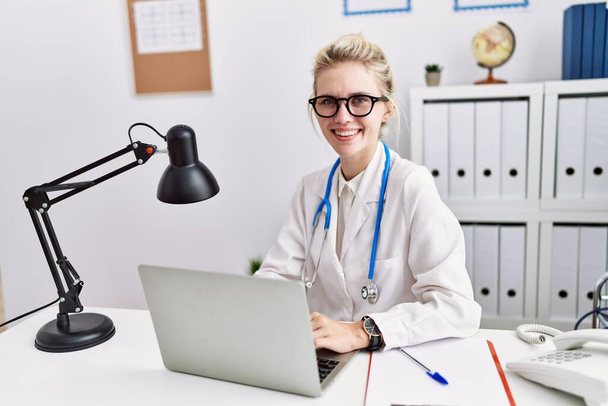Jonge blonde vrouw arts met behulp van laptop werken in kliniek - Foto, afbeelding