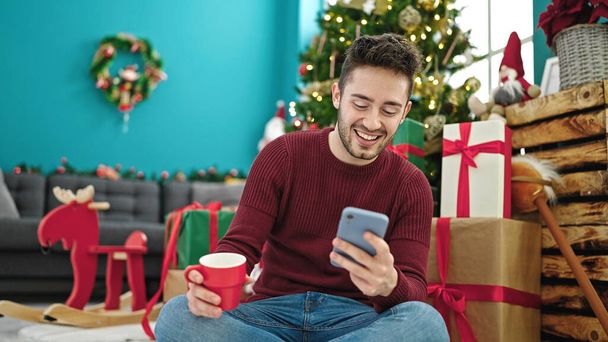 Giovane uomo ispanico utilizzando smartphone bere caffè celebrando il Natale a casa - Foto, immagini