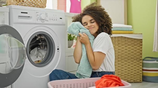 Young beautiful hispanic woman washing clothes touching face with clean towel at laundry room - Photo, Image