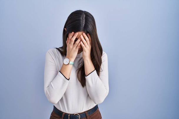 Молодая брюнетка, стоящая на синем фоне с грустным лицом, закрывающим лицо руками во время плача. депрессия.  - Фото, изображение
