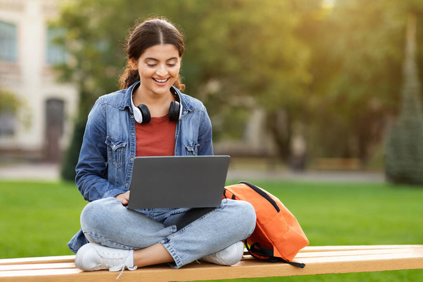 Een glimlachende indiaanse studente die op een laptop zit, zich voorbereidt op examens in de buitenlucht, rust neemt op de universiteitscampus. Technologie, onderwijs en telewerkconcept, kopieerruimte - Foto, afbeelding