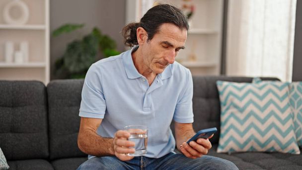Uomo di mezza età in possesso di vetro d'acqua utilizzando smartphone a casa - Foto, immagini