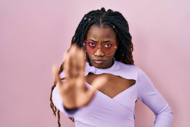 Femme africaine avec les cheveux tressés debout sur fond rose faisant arrêter de chanter avec la paume de la main. expression d'avertissement avec geste négatif et sérieux sur le visage.  - Photo, image