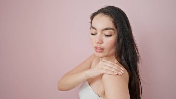 Mujer hispana hermosa joven aplicando tratamiento de loción en el hombro sobre fondo rosa aislado - Foto, Imagen