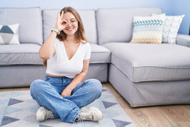 Young caucasian woman sitting on the floor at the living room doing ok gesture with hand smiling, eye looking through fingers with happy face.  - Photo, Image
