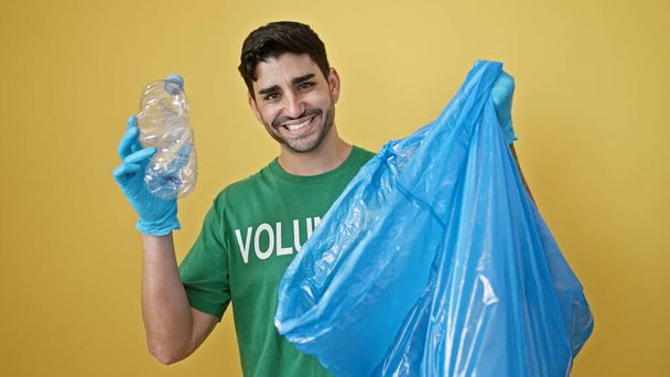 Молодий іспаномовний чоловік доброволець у рукавичках для переробки пластикової пляшки над ізольованим жовтим тлом - Фото, зображення