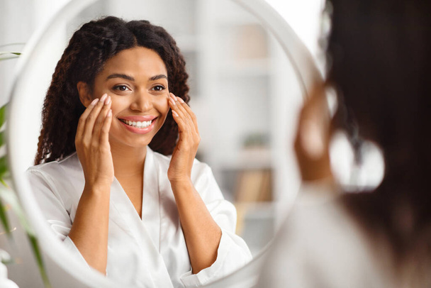 Gyönyörű fekete fiatal nő keres a tükörben, és alkalmazása alatt szem krém, mosolygós vonzó afro-amerikai nő visel fehér selyem Robe Making szépség kezelések otthon, szelektív Focus - Fotó, kép