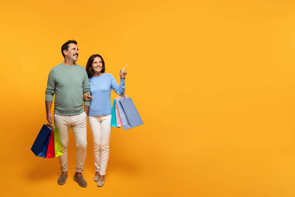 Onnellinen vanhempi eurooppalainen aviomies ja vaimo, jolla on monia pusseja ostoksilla, nauttivat ostoksista, osoittavat sormella vapaata tilaa, eristetty oranssilla studiotaustalla. Suhteet, mainos ja tarjous, huomion myynti - Valokuva, kuva