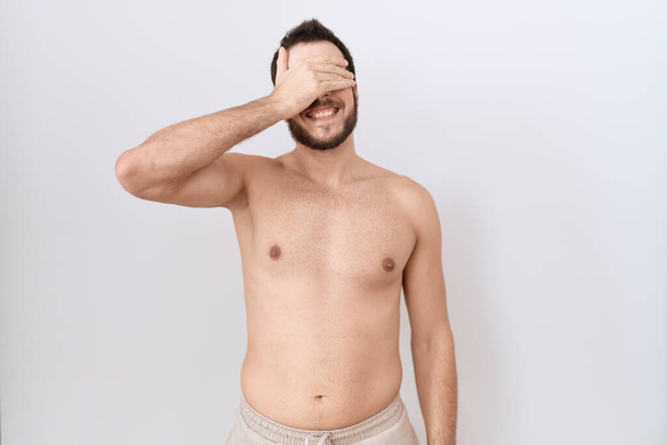 Young hispanic man standing shirtless over white background smiling and laughing with hand on face covering eyes for surprise. blind concept.  - Photo, Image