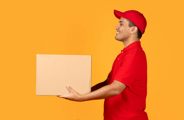 Sivukuva arabi kuriiri kaveri punainen univormu antaa paketti pahvilaatikko sivuun, poseeraa keltainen studio tausta. Moderni toimitus ja nopea toimitus palvelu tarjous - Valokuva, kuva