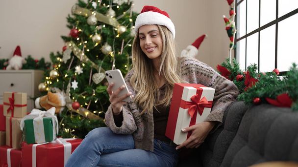 Junge blonde Frau mit Smartphone hält Geschenk zu Hause am Weihnachtsbaum - Foto, Bild