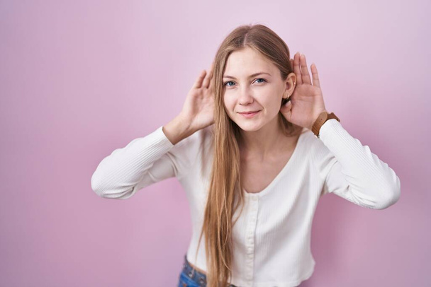 Young caucasian woman standing over pink background trying to hear both hands on ear gesture, curious for gossip. hearing problem, deaf  - Foto, Imagen