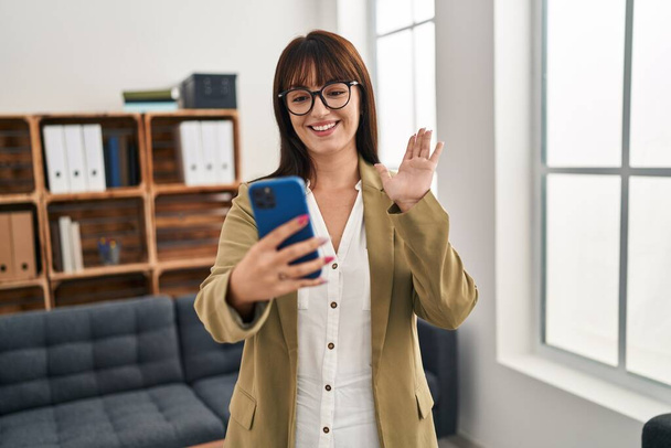 Молодая брюнетка работает в офисе со смартфоном выглядит позитивно и счастливо стоя и улыбаясь с уверенной улыбкой, показывая зубы  - Фото, изображение