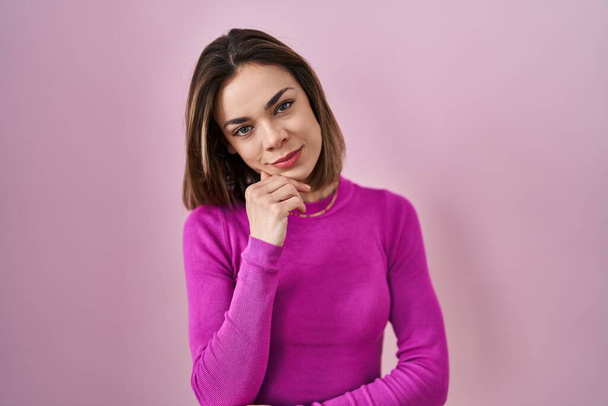 Hispanic woman standing over pink background looking confident at the camera smiling with crossed arms and hand raised on chin. thinking positive.  - Foto, Imagem