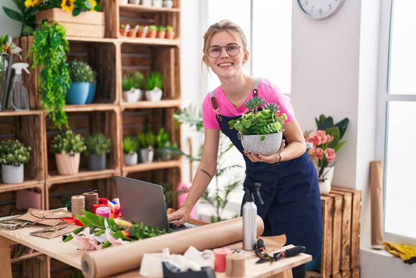 Young blonde woman florist using laptop holding plant at flower shop - Photo, Image