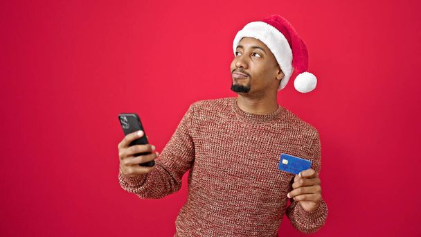 アフリカ系アメリカ人男性 スマートフォンとクレジットカードを使用して,隔離された赤い背景にクリスマスの帽子をかぶります - 写真・画像