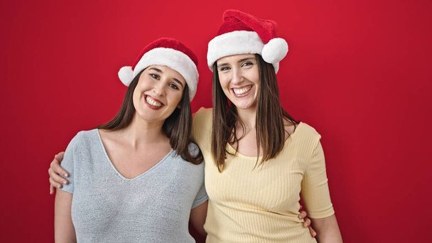 Две женщины обнимают друг друга в рождественской шляпе на изолированном красном фоне - Фото, изображение