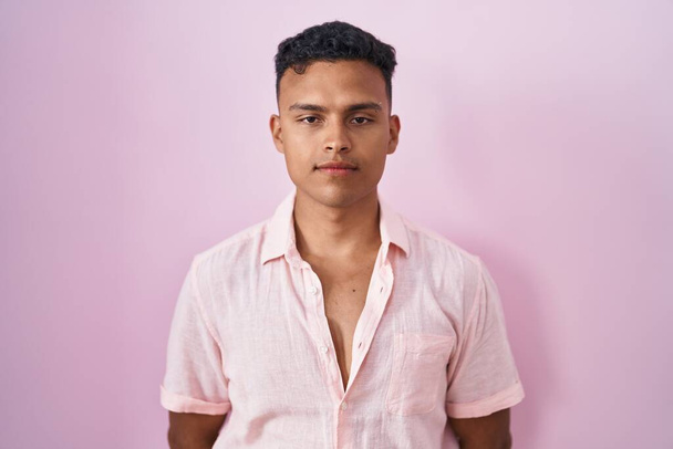 Jonge Spaanse man die over roze achtergrond staat ontspannen met een serieuze uitdrukking op zijn gezicht. eenvoudig en natuurlijk kijkend naar de camera.  - Foto, afbeelding
