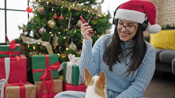 Jonge Spaanse vrouw met chihuahua hond die thuis naar muziek luistert met koptelefoon en kerstmuts op - Foto, afbeelding