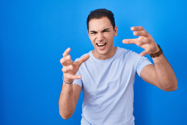 Fiatal spanyol férfi áll a kék háttér felett kiabálva frusztrált a düh, kezek próbál fojtogatni, kiabálva őrült  - Fotó, kép