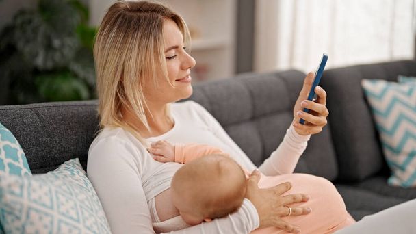 Moeder en dochter zitten op de bank borstvoeding baby met behulp van smartphone thuis - Foto, afbeelding