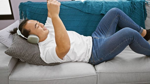 Joyful young chinese man lying on the sofa, confidently listening and dancing to music at home, immersed in the audio world with headphones, expressing positive lifestyle and happiness. - Foto, afbeelding