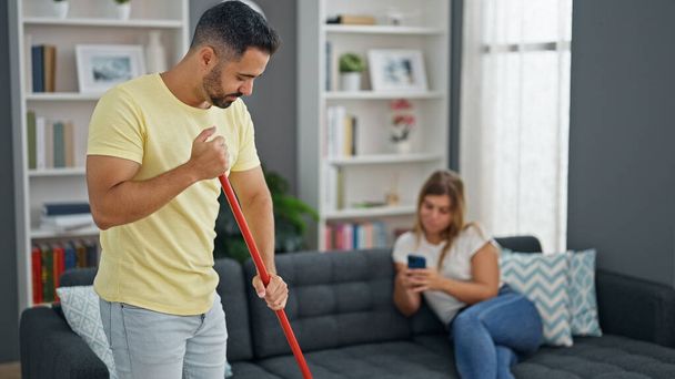 Mann und Frau machen Hausarbeit, während Freundin zu Hause Smartphone nutzt - Foto, Bild