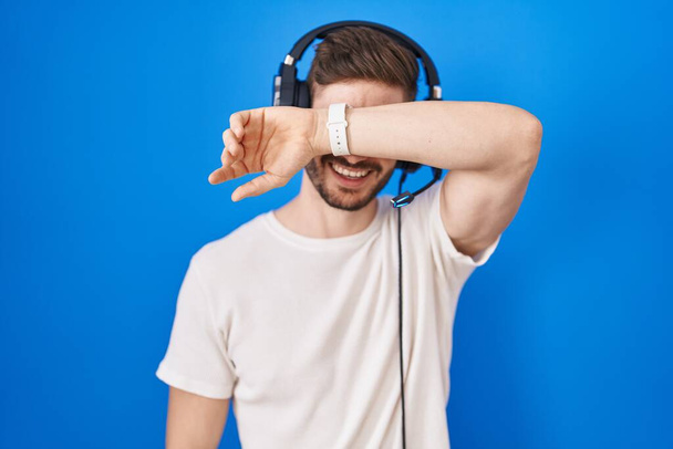 Ισπανόφωνος άνδρας με γενειάδα ακούγοντας μουσική φορώντας ακουστικά που καλύπτουν τα μάτια με το χέρι χαμογελώντας χαρούμενα και αστεία. τυφλή έννοια.  - Φωτογραφία, εικόνα