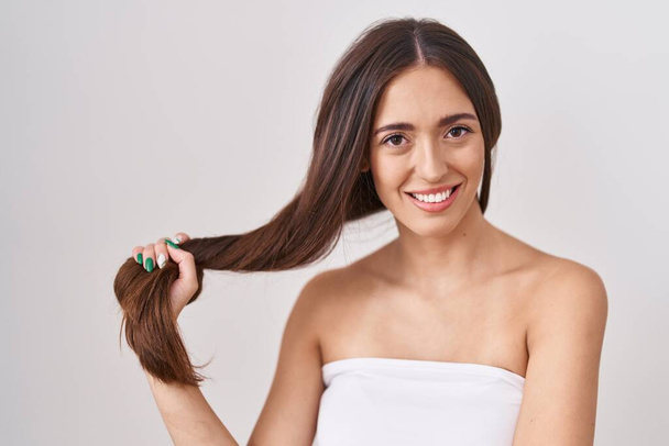 Νεαρή Ισπανίδα που κρατάει τα μαλλιά της με το χέρι της να δείχνει θετική και χαρούμενη να στέκεται και να χαμογελάει με αυτοπεποίθηση να δείχνει τα δόντια της  - Φωτογραφία, εικόνα