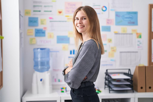 Молодая блондинка бизнес-работница улыбается уверенно стоя со скрещенными руками в офисе - Фото, изображение
