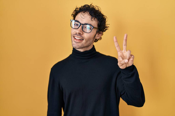 Hispanic man standing over yellow background showing and pointing up with fingers number two while smiling confident and happy.  - Foto, afbeelding