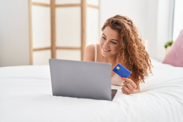 Молода красива іспаномовна жінка використовує ноутбук і кредитну картку, сидячи на ліжку в спальні
 - Фото, зображення